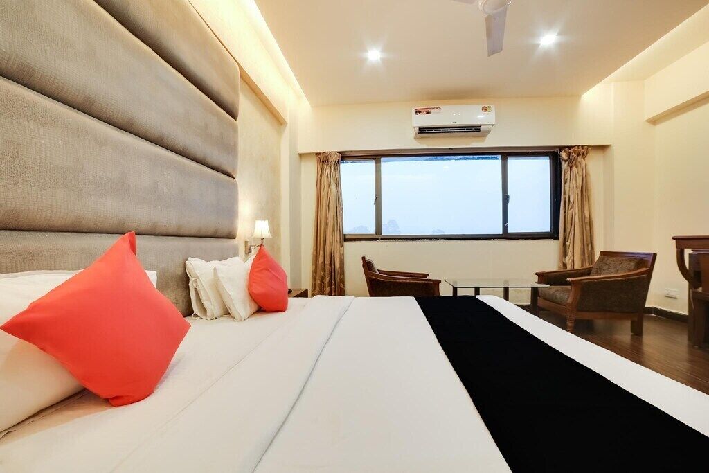 A hotel room with a large bed, a desk, and a chair. There is an air conditioning unit on the wall and a window with curtains.