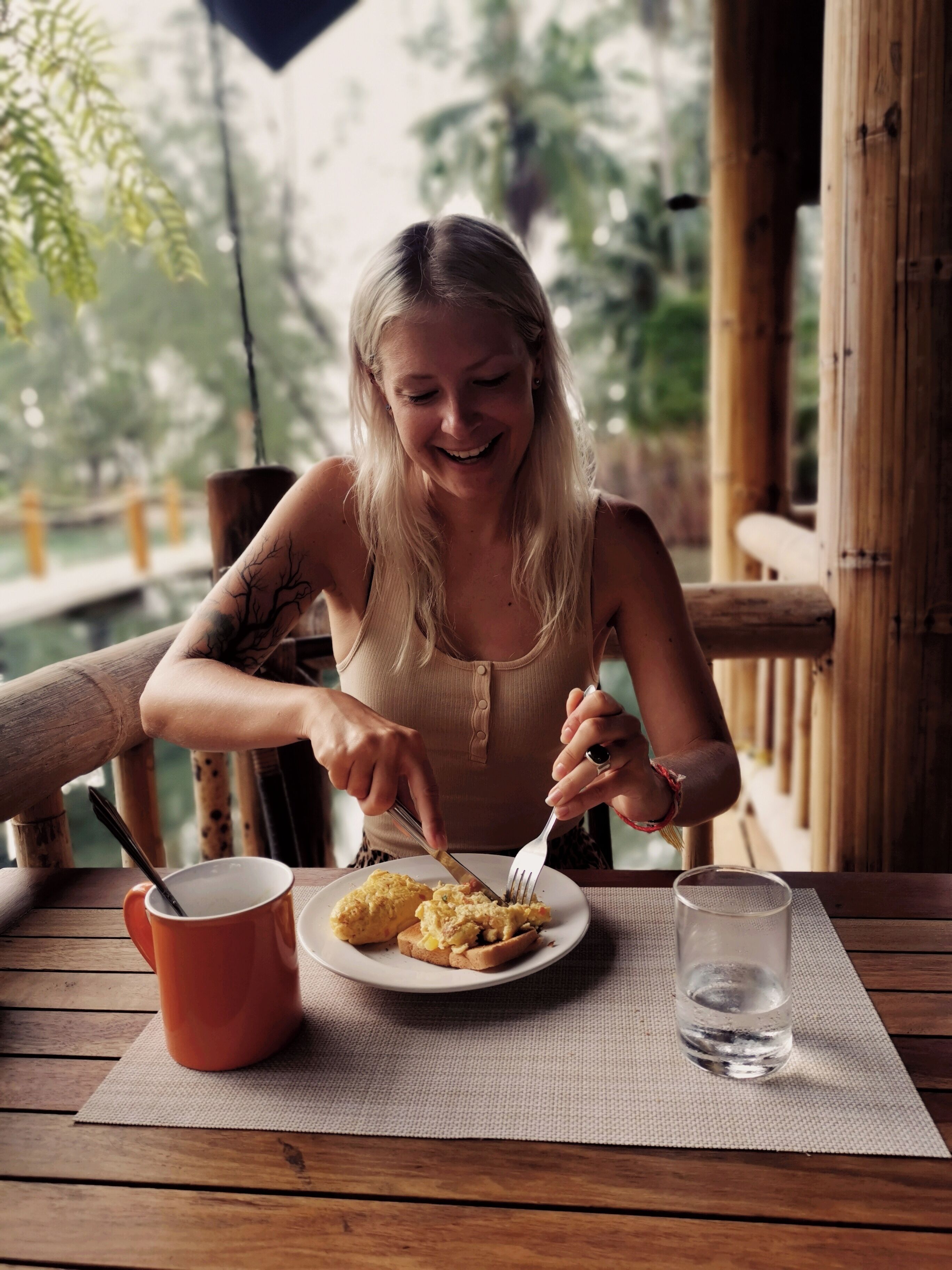 Una persona cenando en una mesa de madera al aire libre, con un plato de comida, un vaso de agua y una taza roja.