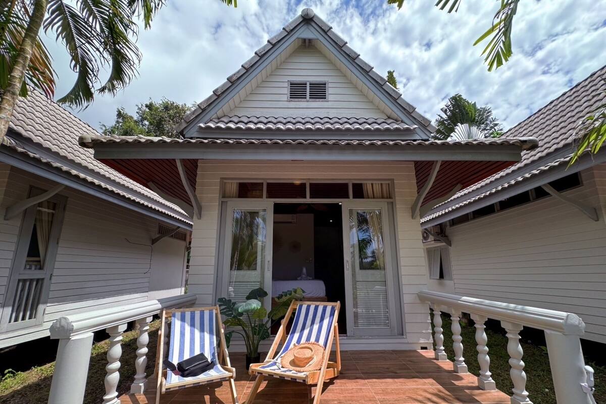A two-story house with a tiled porch, two lounge chairs, and a small garden area.