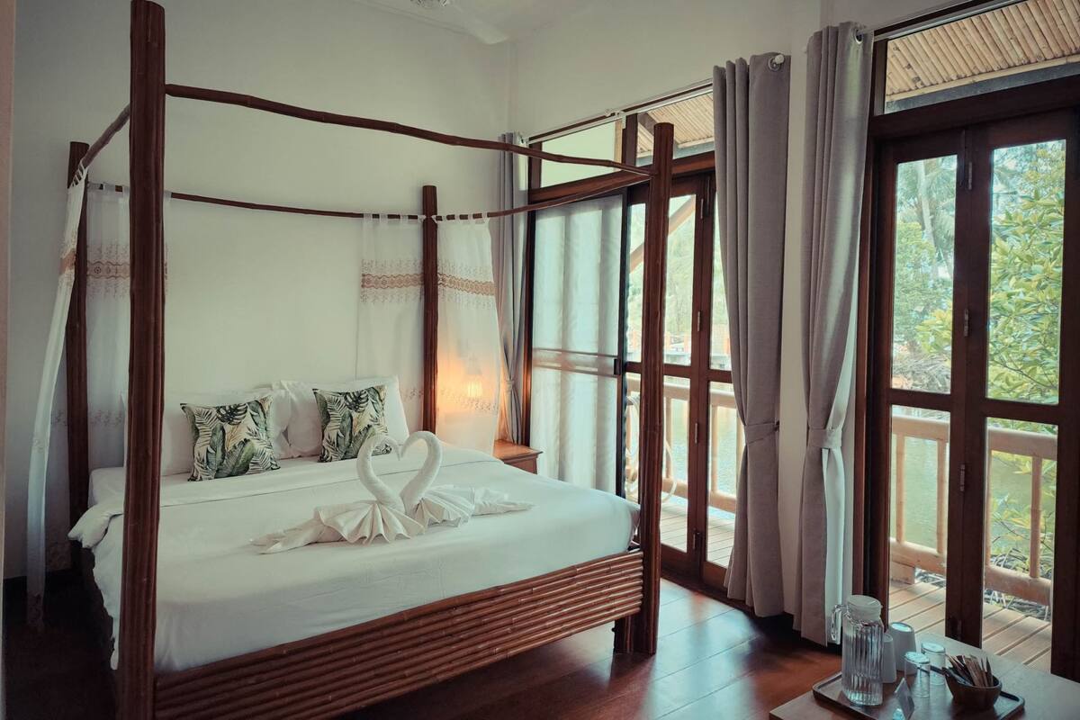 A four-poster bed with a swan-shaped bedspread, a ceiling fan, and a view of a wooden deck through large windows.