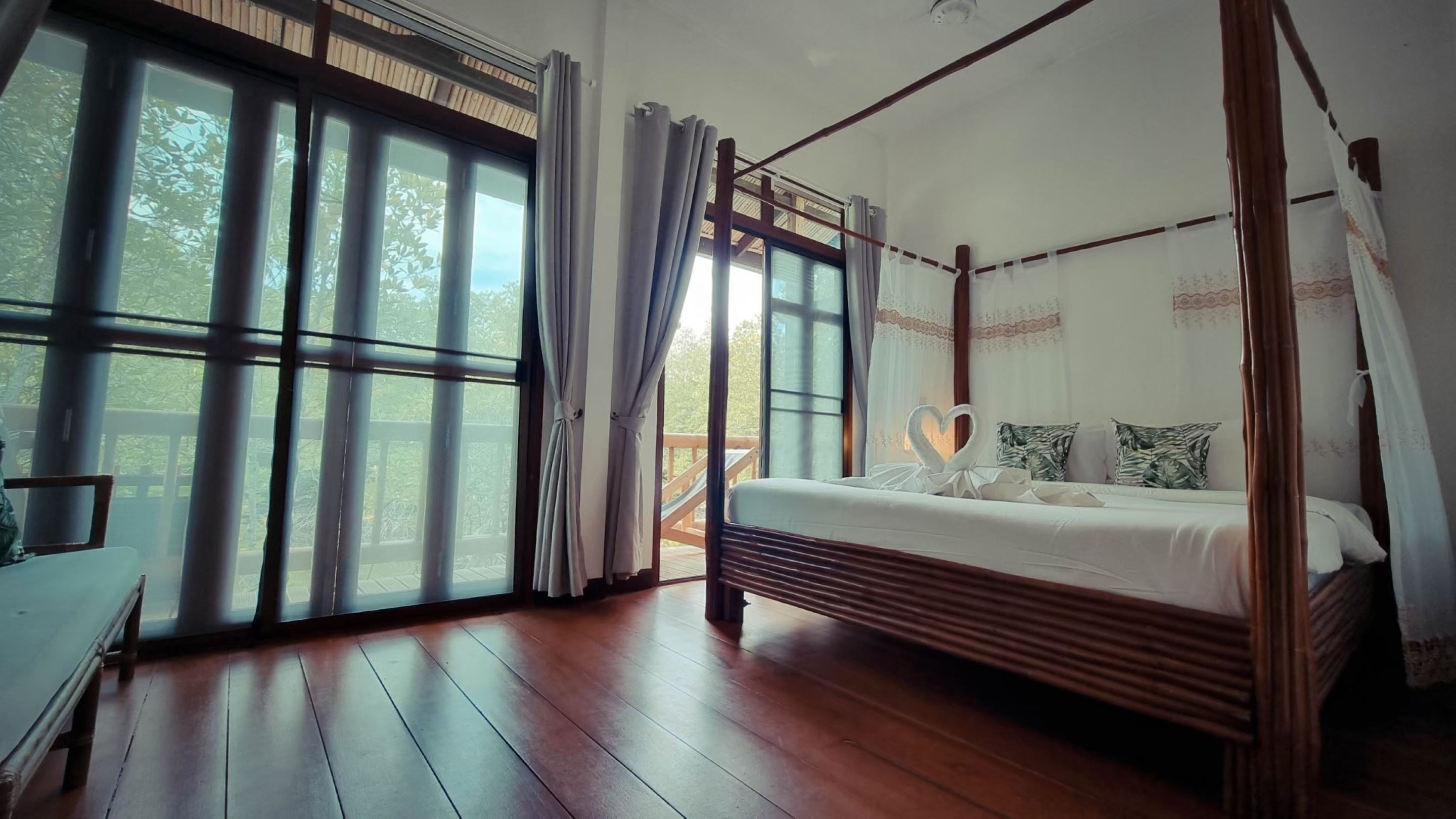 A four-poster bed with a wooden frame and white bedding, large windows with curtains, and a wooden floor.