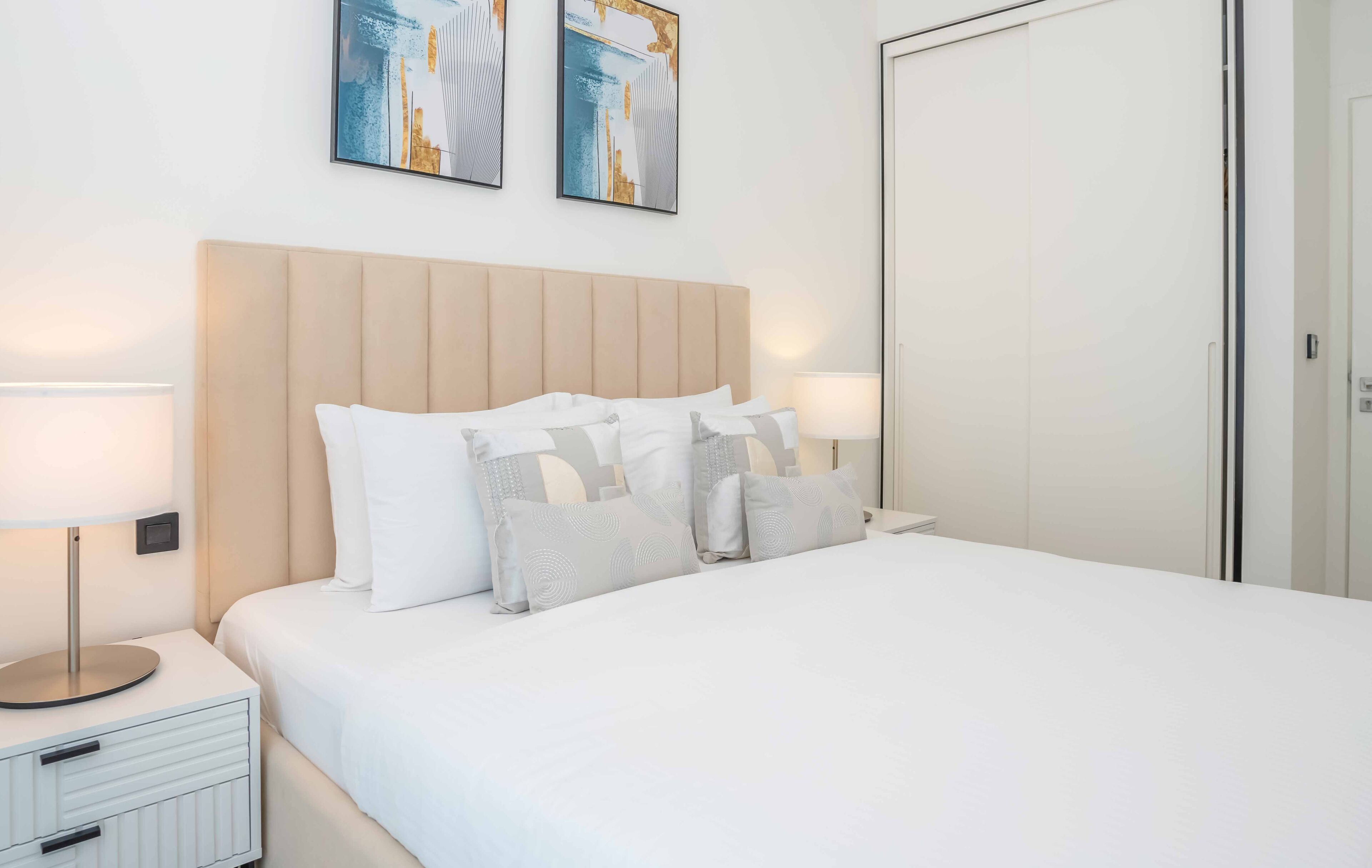 Un lit bien fait, avec du linge de lit blanc et plusieurs oreillers, une table de chevet avec une lampe, et deux tableaux encadrés accrochés au mur.