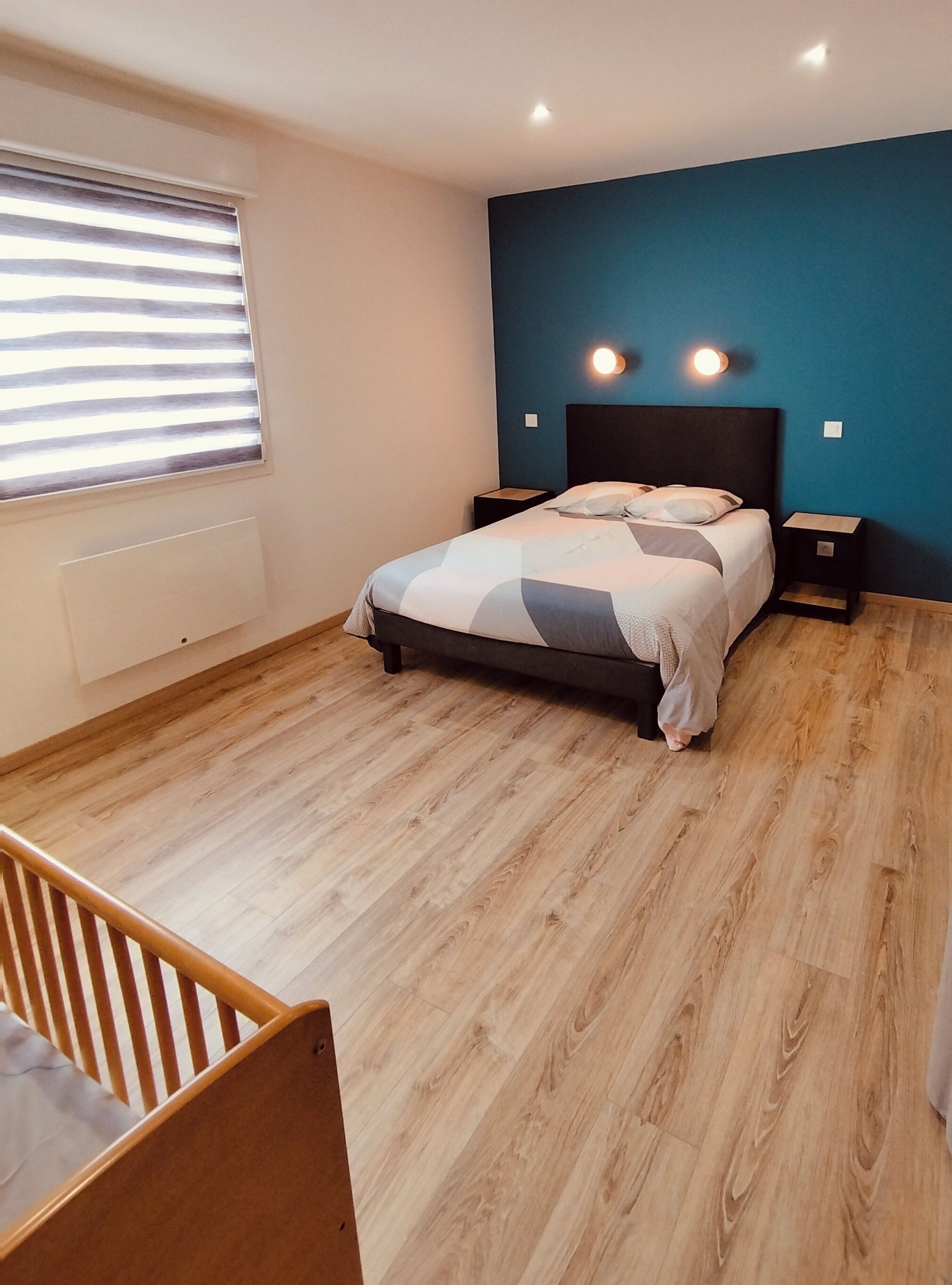 Une chambre à coucher comprenant un lit, deux tables de chevet, une fenêtre avec des stores, un radiateur et un plancher en bois.
