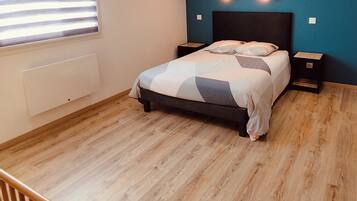 Une chambre à coucher comprenant un lit, deux tables de chevet, une fenêtre avec des stores, un radiateur et un plancher en bois.