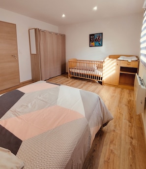 Une chambre avec un plancher en bois, un lit recouvert d’une couvre-lit à motifs, un berceau en bois, une armoire, un bureau et une œuvre d’art accrochée au mur.