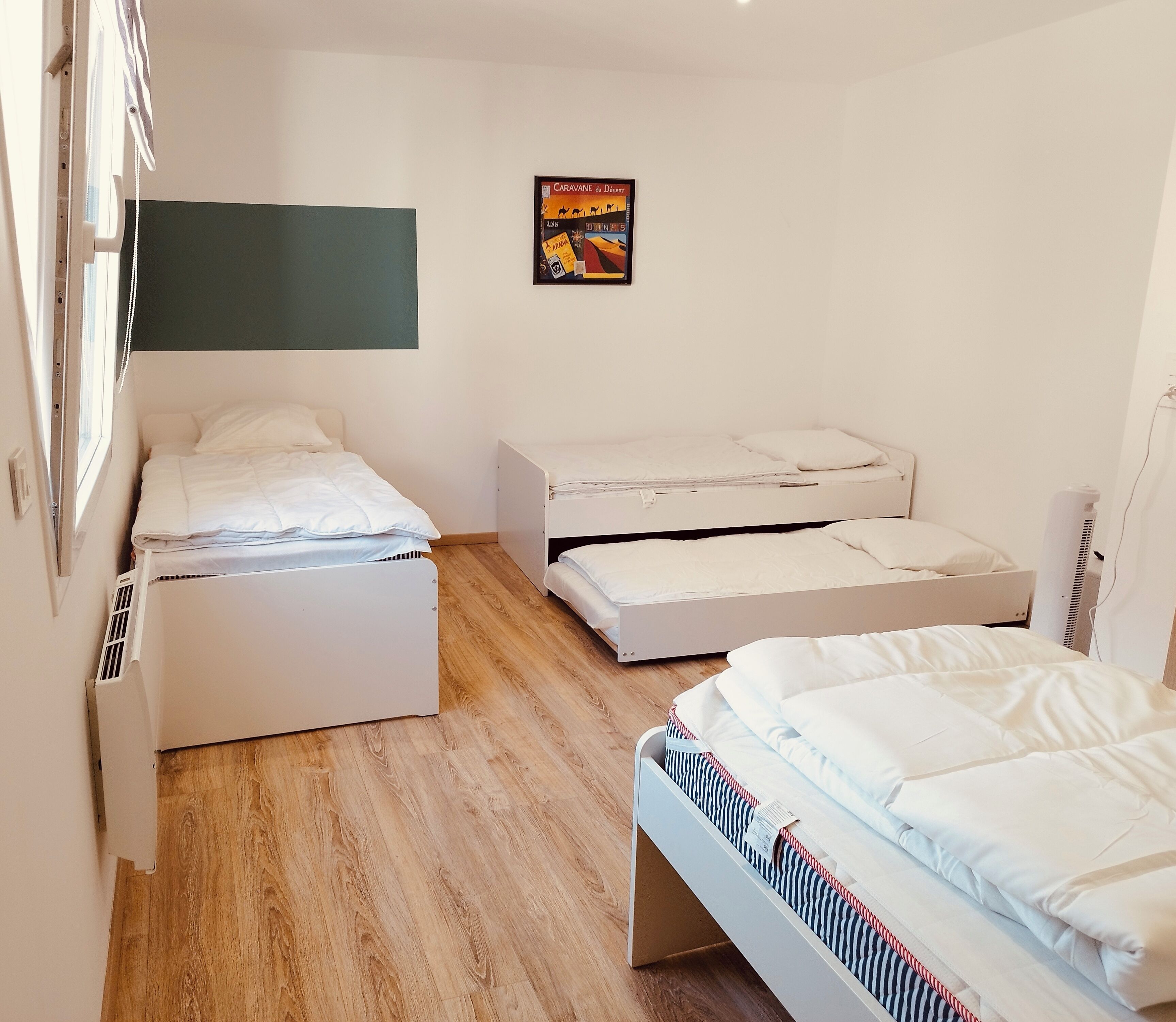 Ein Zimmer mit drei Betten, einem Fenster und einem Wandbild.