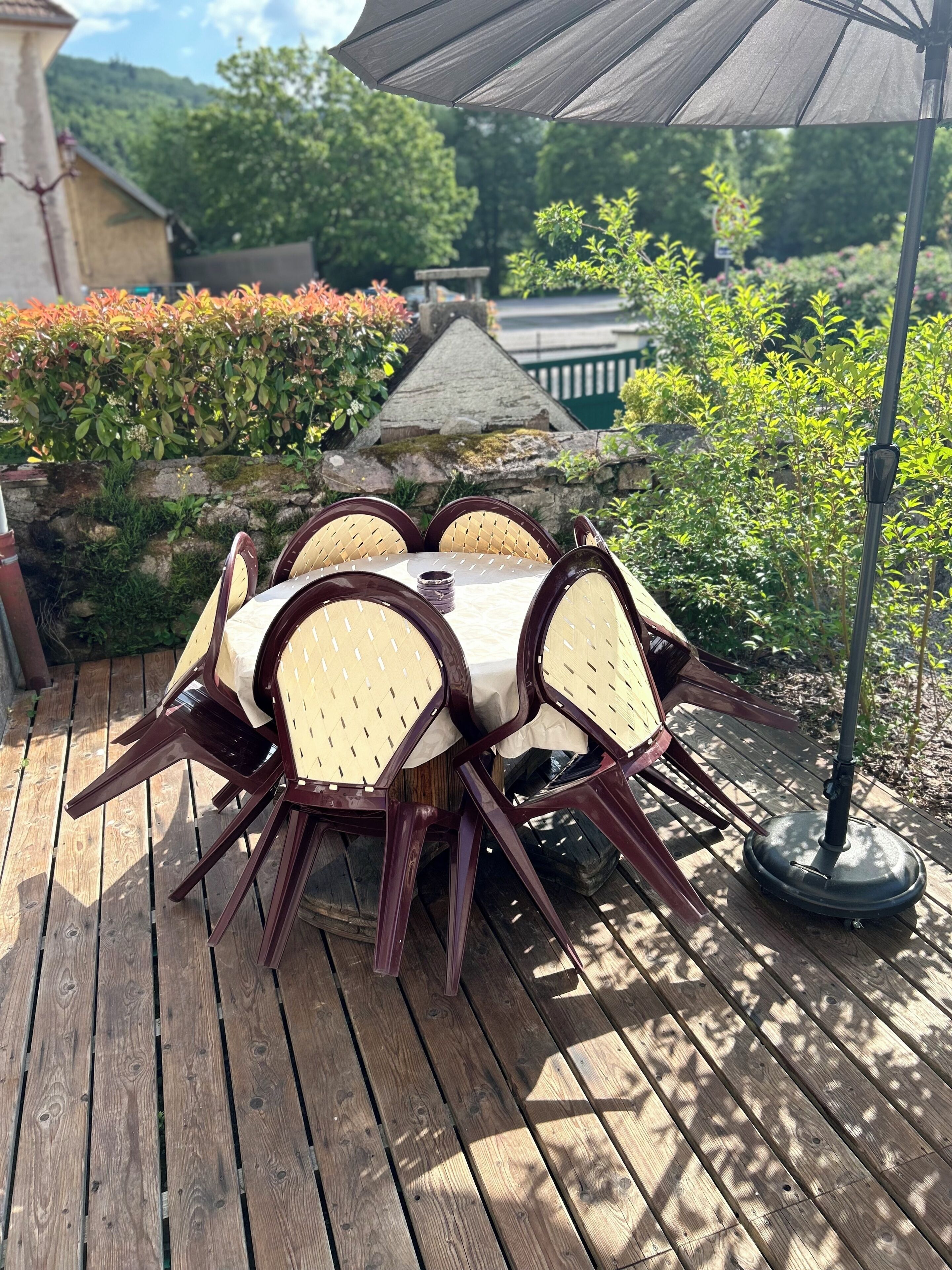 Eine Holzterrasse mit einem runden Tisch, Stühlen und einem Sonnenschirm, im Hintergrund eine Steinmauer.