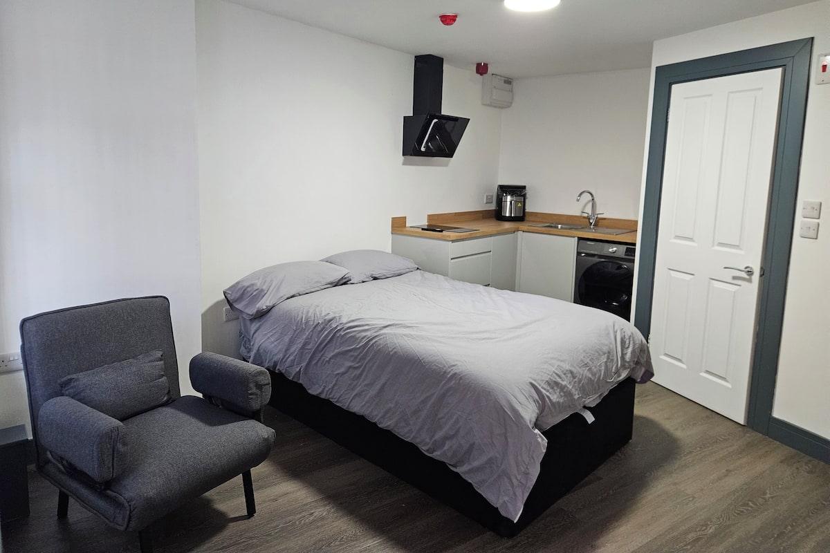 A compact room with a bed, a chair, a small kitchenette, and a door.