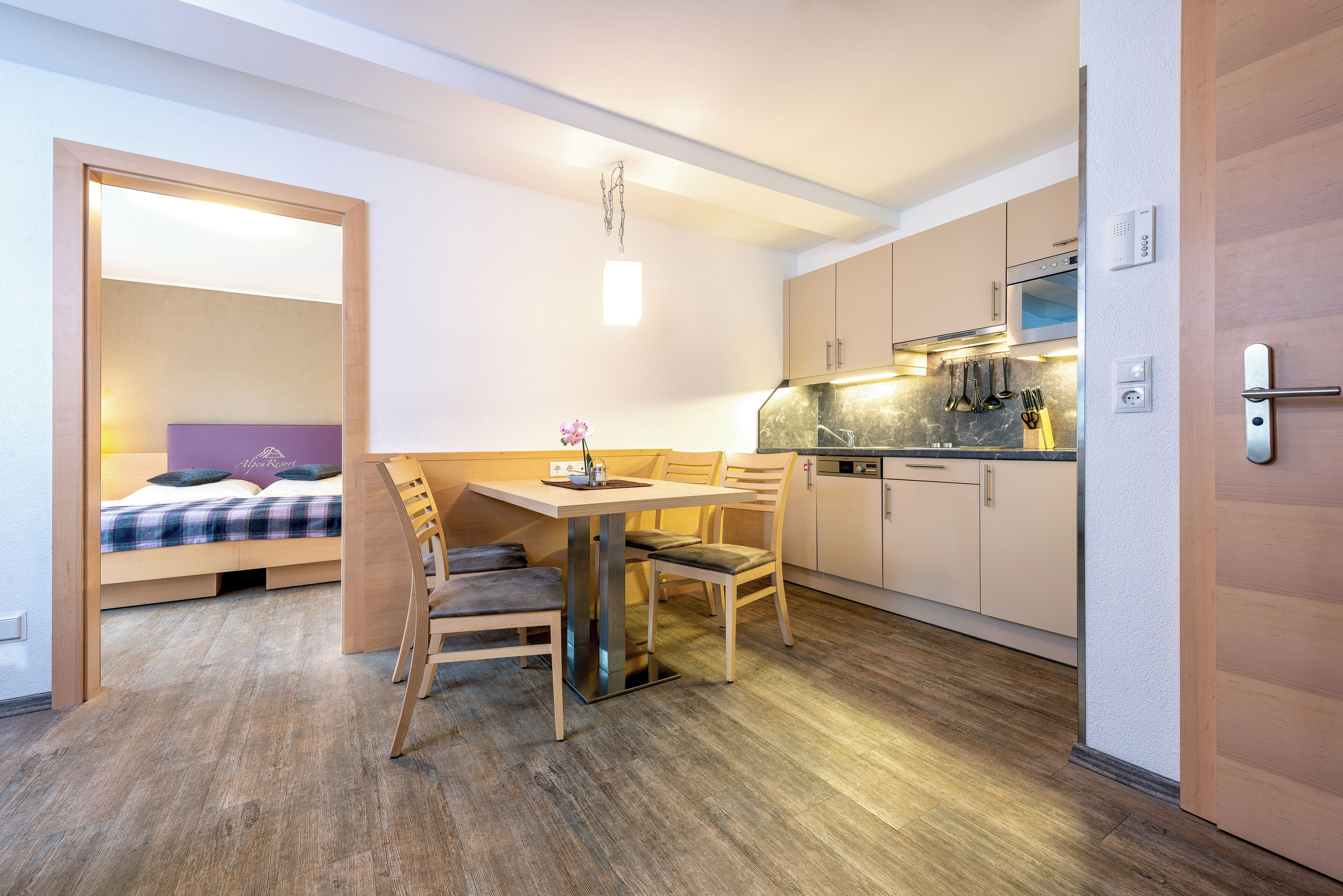 Una habitación de hotel compacta con kitchenette, un comedor con mesa y sillas, y una cama con cabecera morada.
