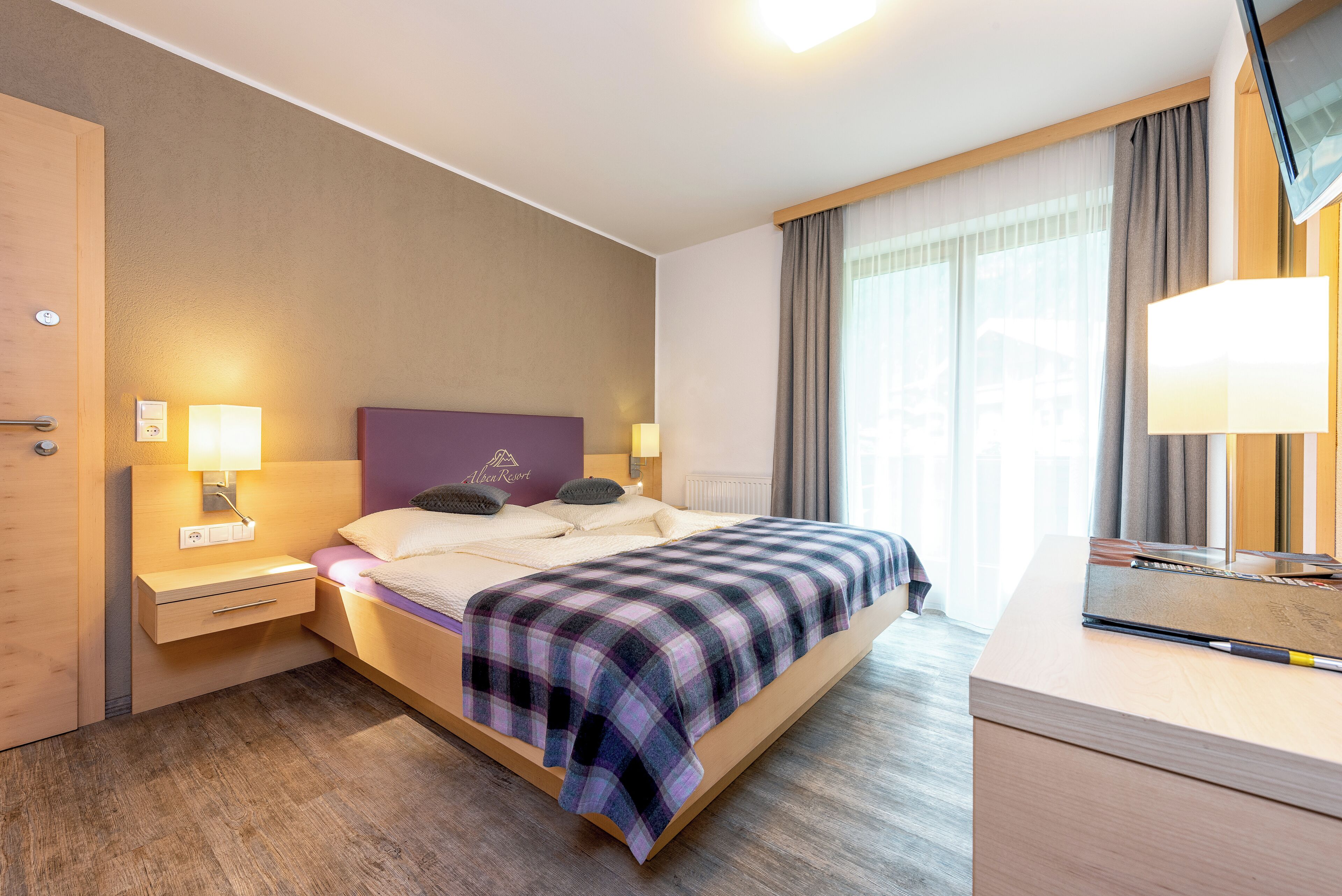 A modern hotel room with a large bed, bedside lamps, a wooden nightstand, a desk with a lamp, and a view of the outdoors through the window.