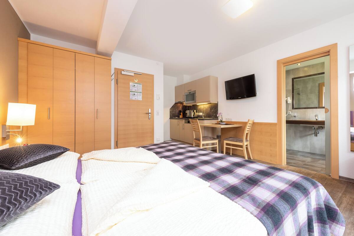 A hotel room with a bed, a desk, a chair, a TV, and a shower area.