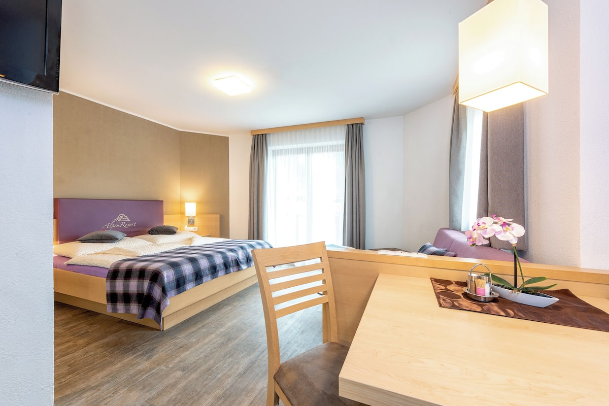 A hotel room with a bed, a desk, a chair, a lamp, and a window with curtains.