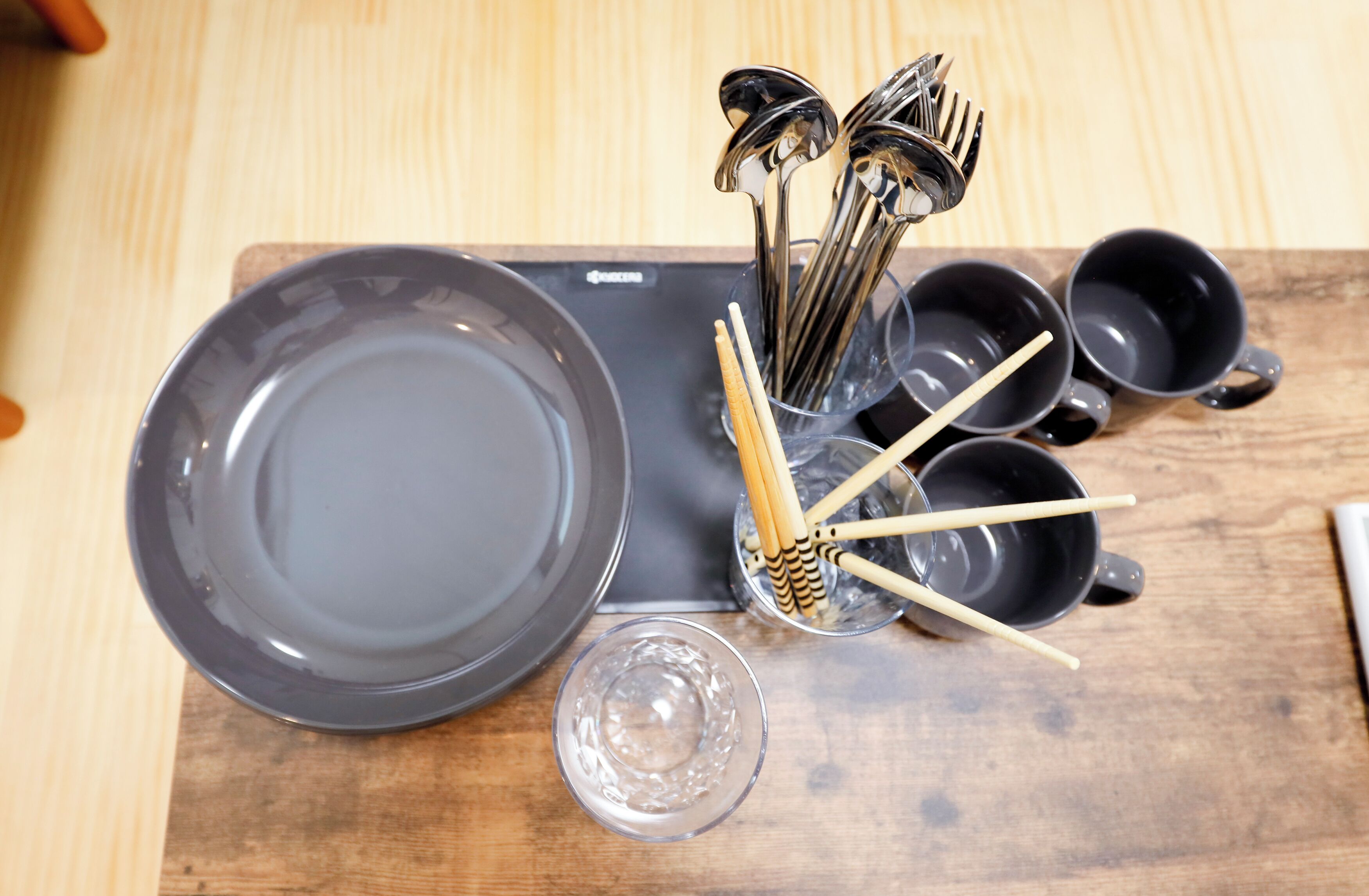 A wooden table with a black tray holding a clear glass, a black bowl, and a set of chopsticks and spoons in a clear holder.