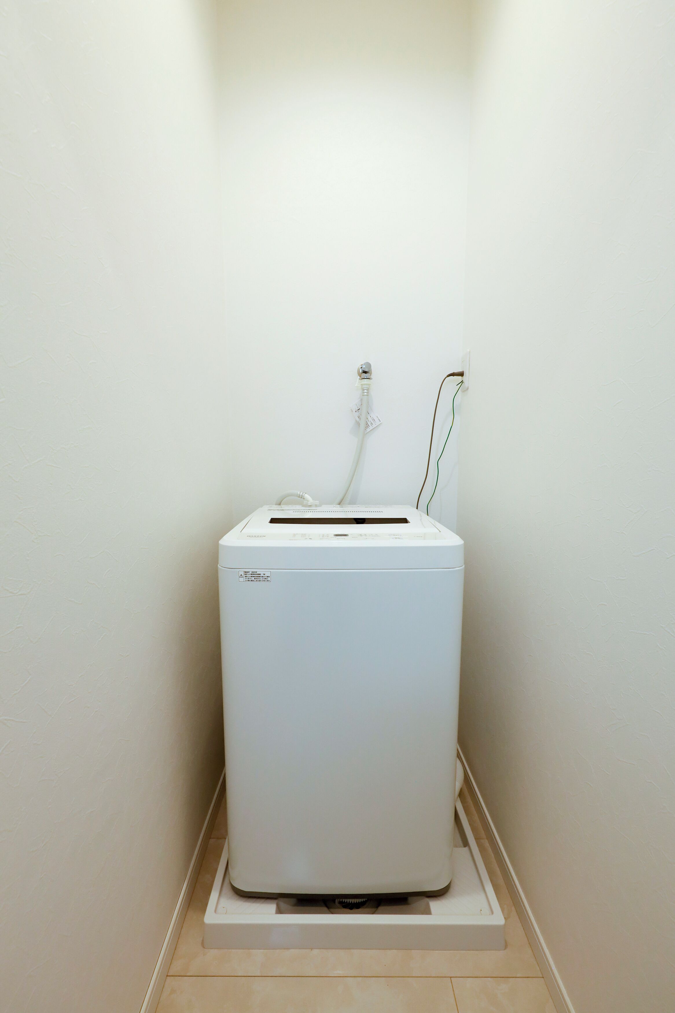 A white, compact, wall-mounted laundry machine in a corner with a power outlet and cables.