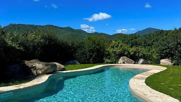 A clear blue pool with a stone border, surrounded by lush greenery and mountains in the background. - Luna & Limoni Country (Muravera)
