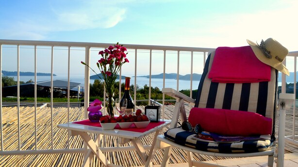 A balcony with a white railing, a striped chair with a red cushion, a small table with a bottle, glasses, and a bowl of fruit.
