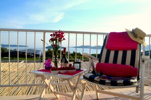 A balcony with a white railing, a striped chair with a red cushion, a small table with a bottle, glasses, and a bowl of fruit.