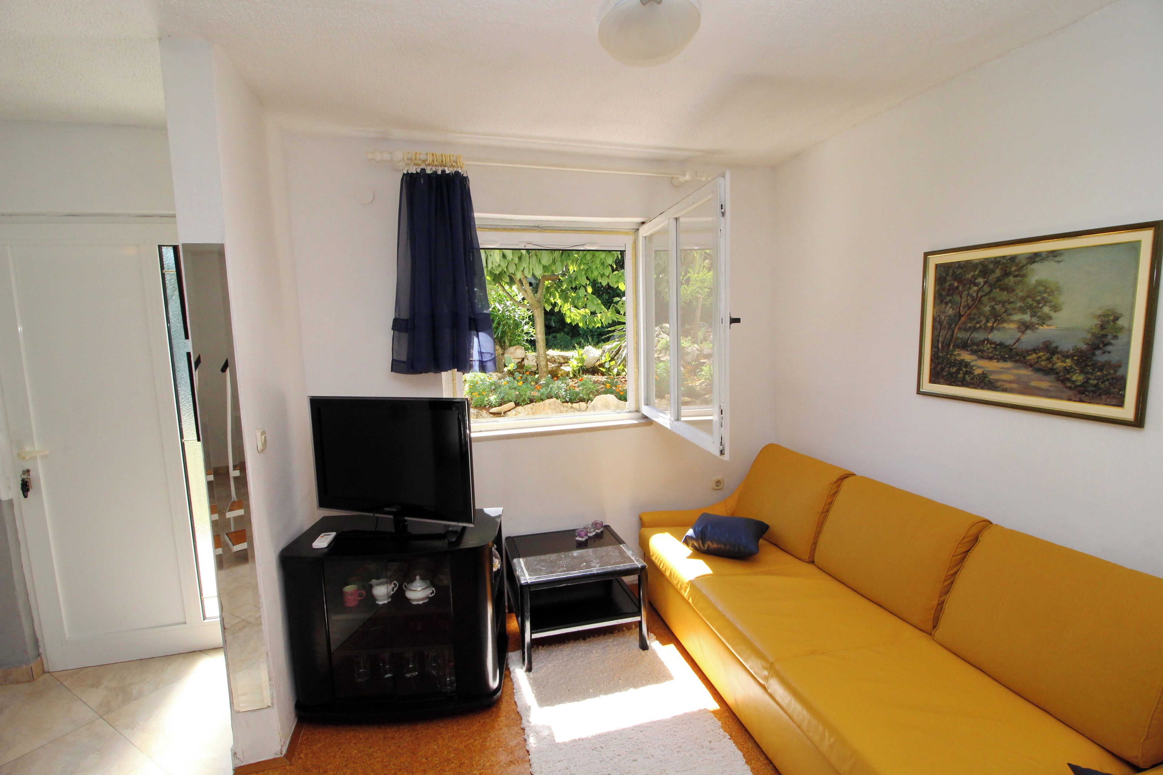 A living room with a yellow sofa, a TV on a stand, a small table, and a window with curtains.