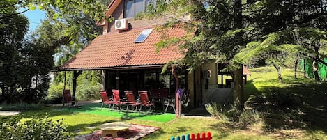 A house with a red-tiled roof, a covered patio with red chairs, and a grassy yard with colorful play equipment.