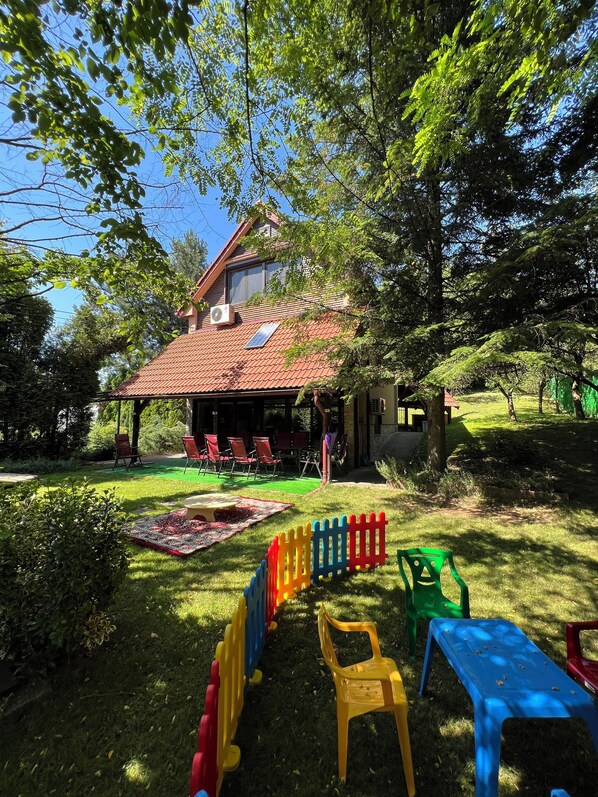 Ein Haus mit rotem Ziegeldach, einer überdachten Terrasse mit roten Stühlen und einem grünen Garten mit bunten Spielgeräten.