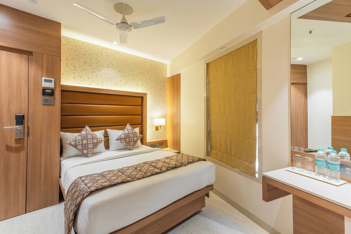 Habitación de hotel con una cama grande, una mesita de noche con botellas de agua, una lámpara y un ventilador de techo, además de un amplio ventanal con vistas.