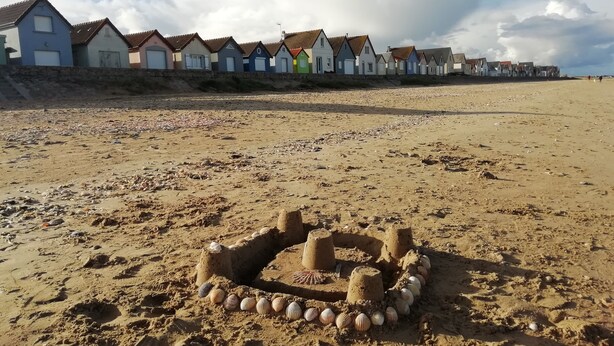 A sandy beach with a sandcastle in the foreground and colorful houses in the background. - G2 LA Cour à Proximité de la Plage de Ravenoville Plage (Sainte-Mère-Église)