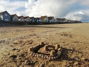 Une plage de sable avec un château de sable au premier plan et des maisons colorées en arrière-plan.