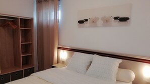 A neatly made bed with white bedding, a wooden wardrobe, and a wall-mounted shelf with decorative items. - G2 LA Cour à Proximité de la Plage de Ravenoville Plage (Sainte-Mère-Église)