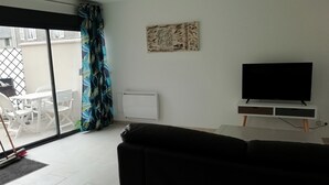 A living room with a black sofa, a TV on a stand, a dining table with chairs, and a sliding glass door leading to an outdoor area. - G2 LA Cour à Proximité de la Plage de Ravenoville Plage (Sainte-Mère-Église)