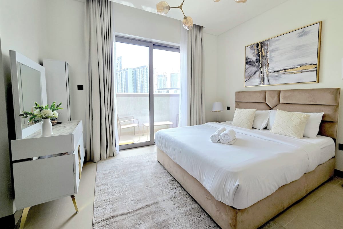 A modern hotel room with a large bed, a desk with a vase of flowers, a large window with curtains, and a city view.