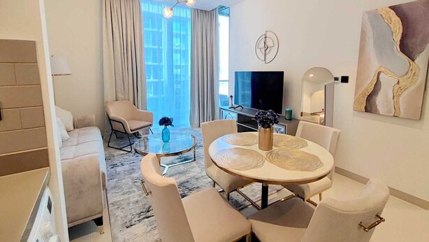 Een moderne woonkamer met een ronde eettafel, beige banken, een televisie en een groot raam met gordijnen.