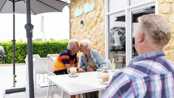 Een buitenruimte met witte stoelen en tafels, een stenen muur met een neonreclame en een glazen deur naar een restaurant.