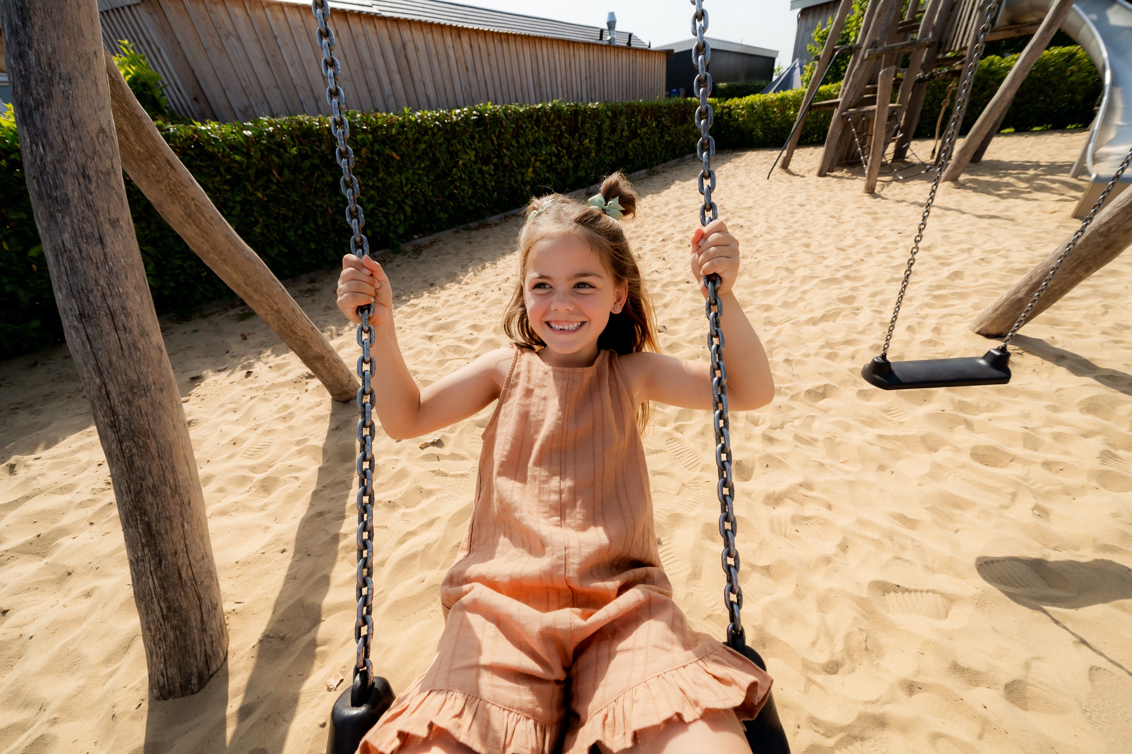 A child on a swing set in a sandy area with a wooden structure and a hedge in the background.
