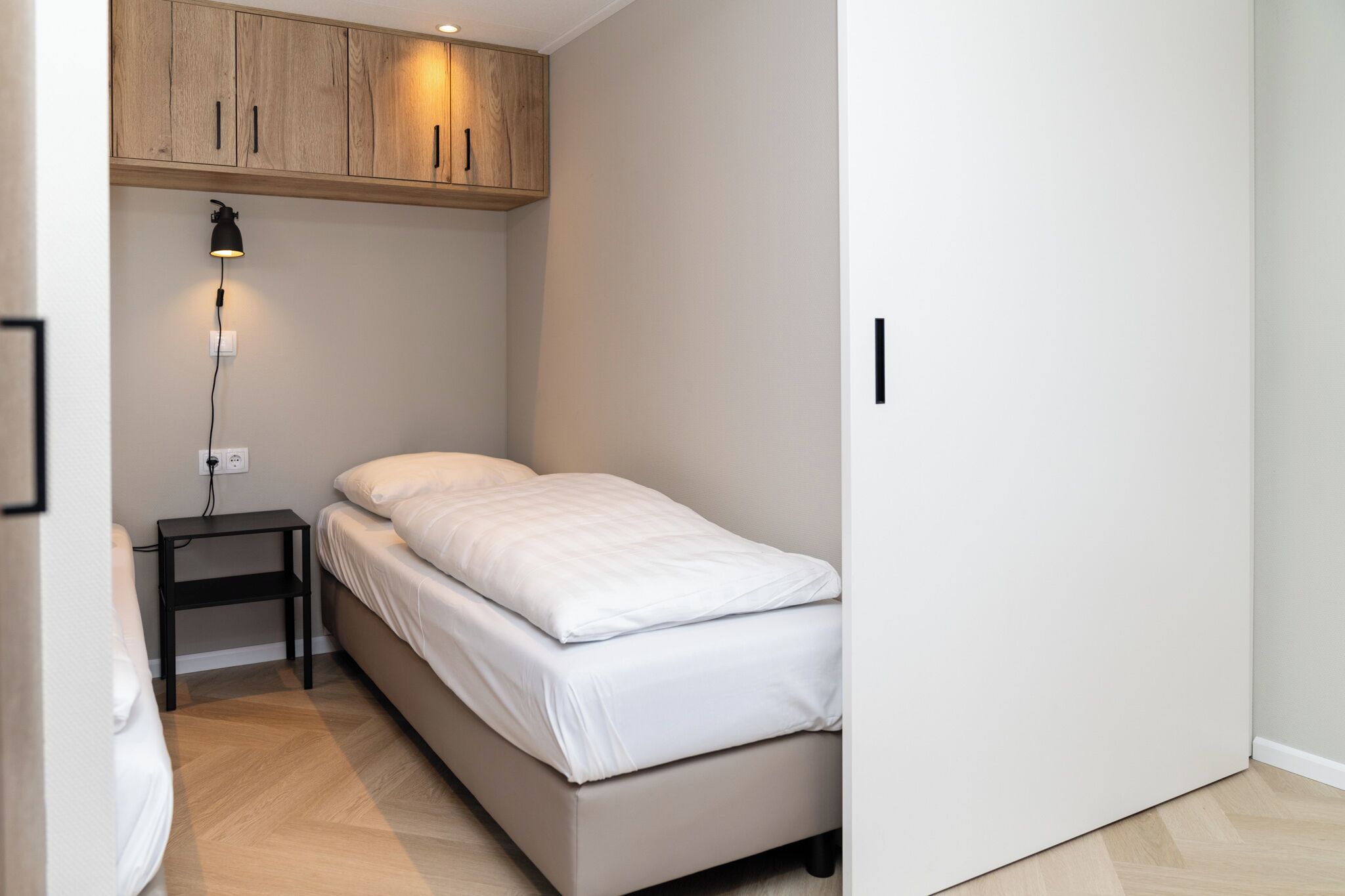 Une petite chambre à coucher, bien rangée, avec un lit simple, une table de chevet, une lampe et des armoires en bois au-dessus.