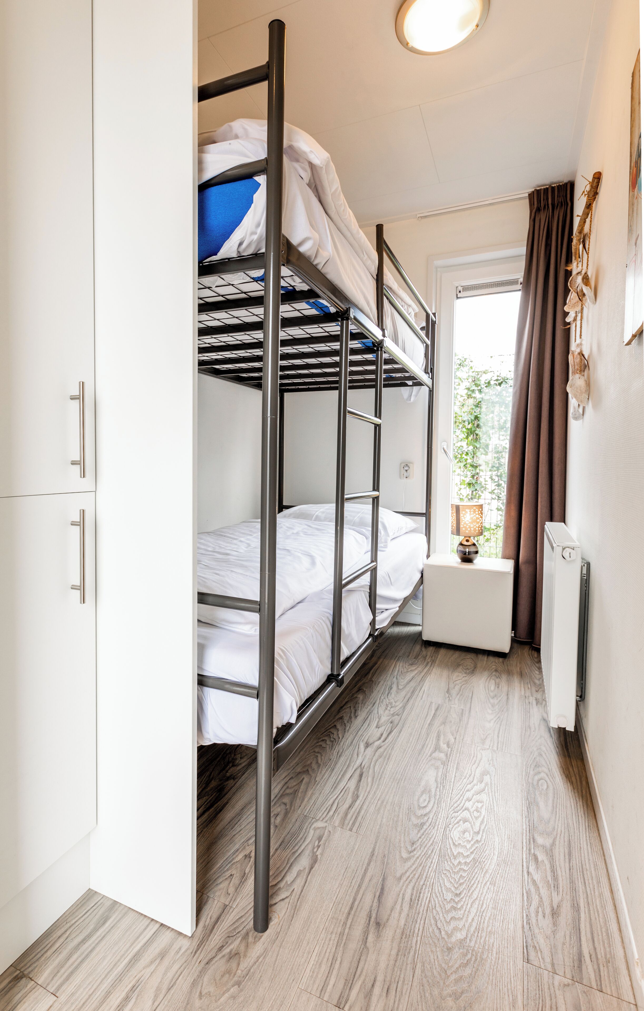 Une petite chambre avec des lits superposés, une petite table de chevet avec une lampe, un radiateur et une fenêtre avec des rideaux.