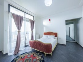 Un dormitorio con una cama grande, una ventana con cortinas, un tapete pequeño y una lámpara roja fijada a la pared.