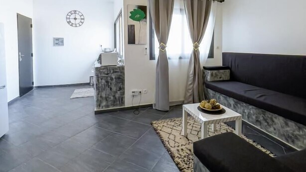 A modern living room with a dark grey tiled floor, a black sofa, a small white table with a bowl of fruit, a grey rug, and a ceiling light fixture. - Kapinga's house (Mbour)
