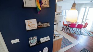 A room with a blue wall, a framed quote, a rainbow flag, and a dining area with orange chairs and a table.