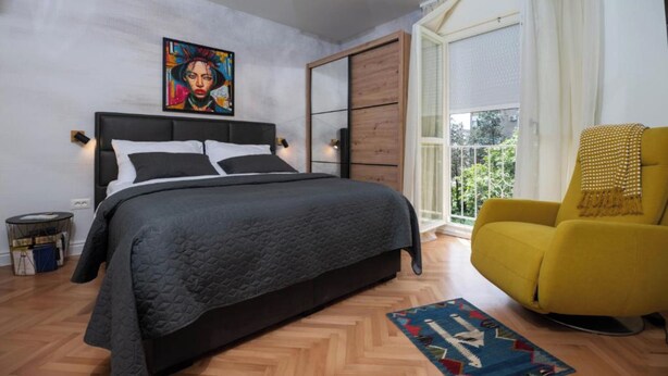 A bedroom with a bed, a yellow armchair, a window with curtains, and a painting on the wall.