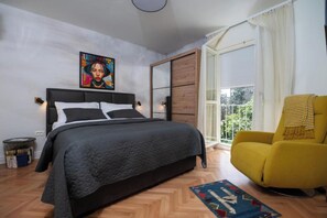 Une chambre à coucher avec un lit, un fauteuil jaune, une fenêtre avec des rideaux et un tableau accroché au mur.