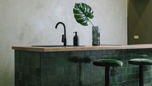 A kitchen with a green tiled backsplash, a wooden countertop, a black faucet, a stainless steel pendant light, and two green bar stools.