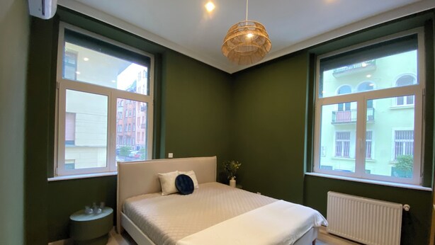 A bedroom with a bed, two windows, a radiator, and a bedside table.
