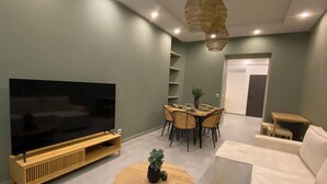 A living room with a flat-screen TV, a round wooden coffee table, a white sofa, a dining table with chairs, and a hanging light fixture.