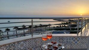 Un balcon avec vue sur la mer et le coucher de soleil, deux verres d’une boisson rouge et un plateau de collations.