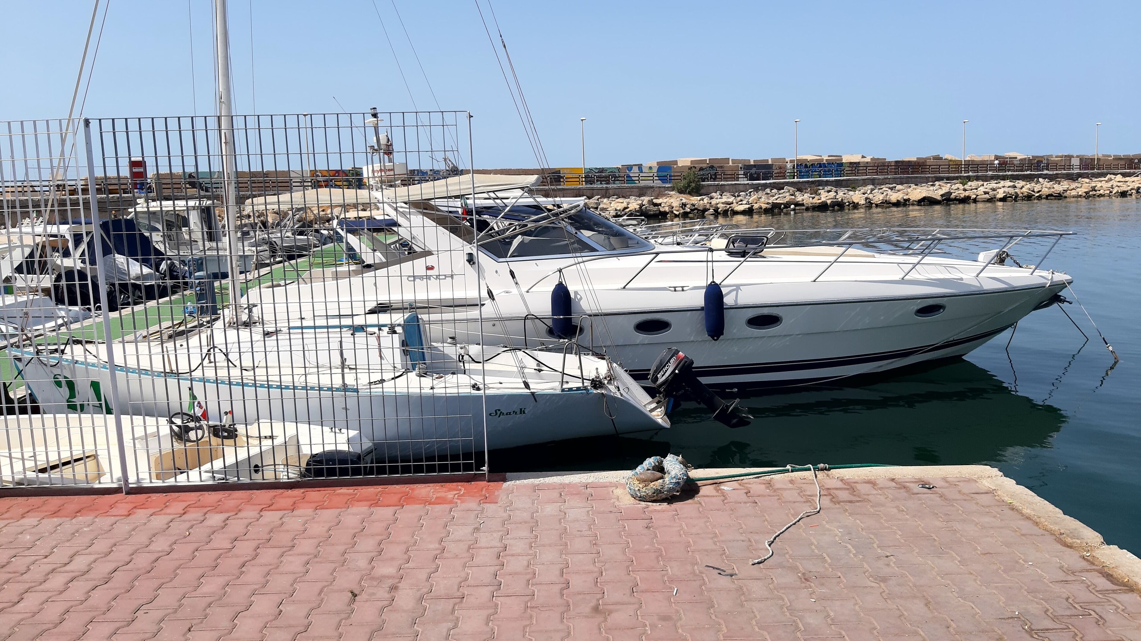 A marina with several boats docked, a paved dock area, and a rocky shoreline in the background.