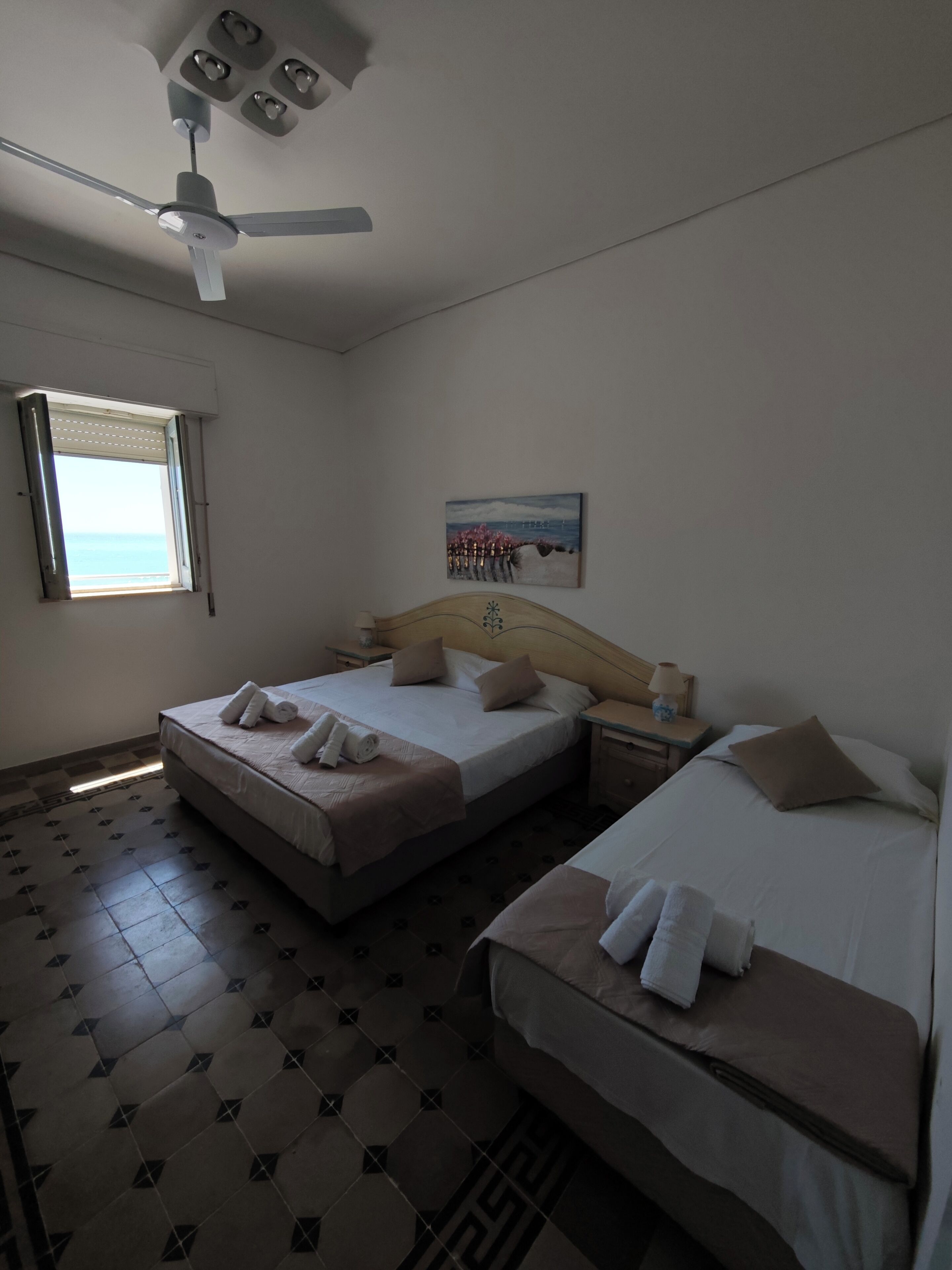 Une chambre d’hôtel avec deux lits, un ventilateur de plafond, une fenêtre avec des stores, un tableau au mur et un sol carrelé.