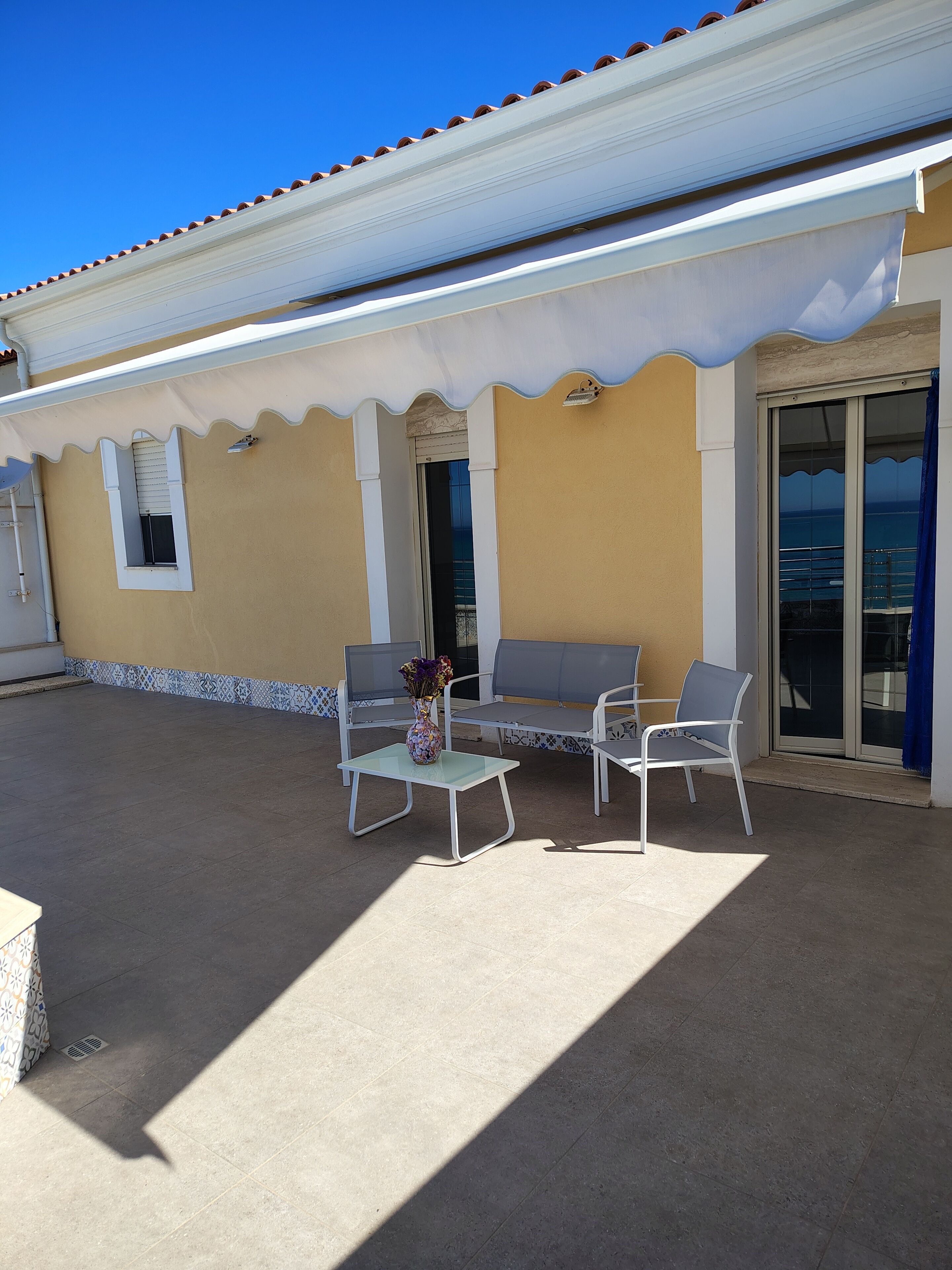 An outdoor patio with a table and two chairs, a vase with flowers, and a view of the sea through a glass door.