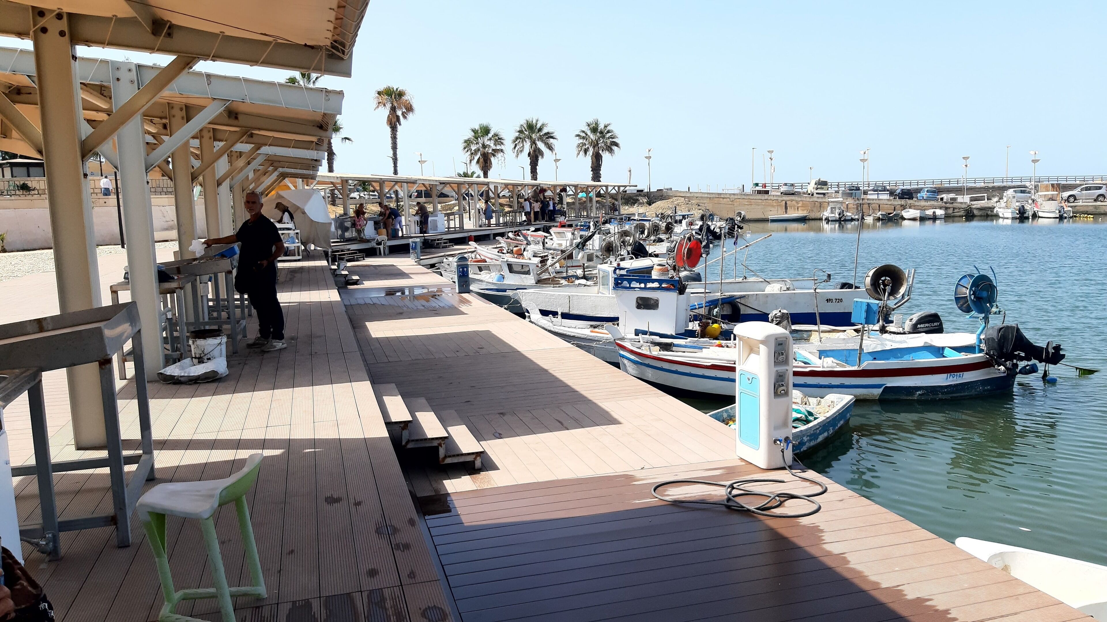 Un puerto deportivo con barcos amarrados, una zona de estar cubierta y un paseo pavimentado.