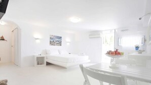 Una habitación blanca con una cama, una mesita con fruta y una silla blanca.
