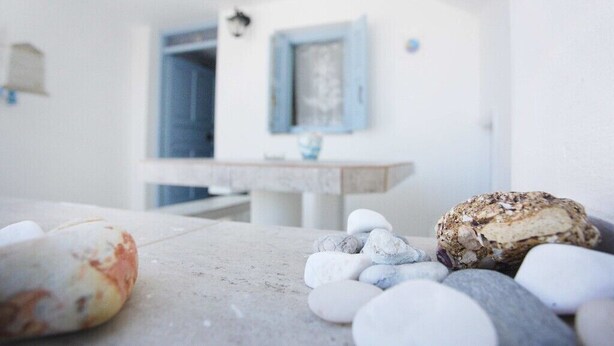 Una mesa de piedra con un pan y guijarros, una pared blanca y un ventanal con marco azul.